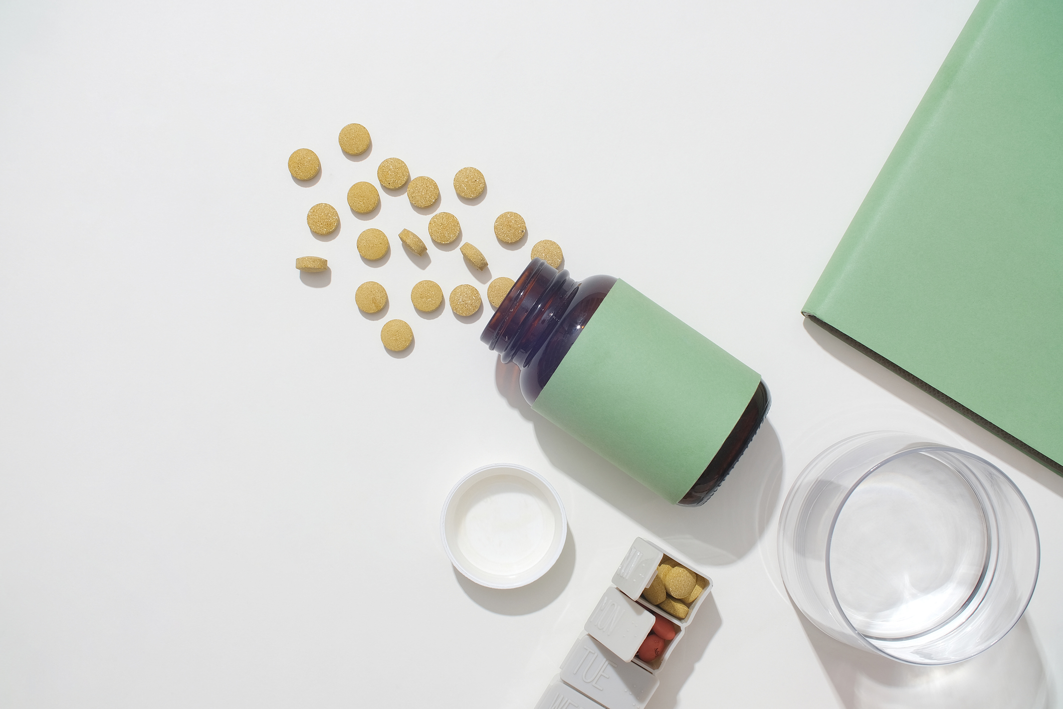 Top view of yellow tablets poured onto the table from an unbranded medicine bottle, a glass of water, a daily pill box and a notebook on a white background. Drug advertising.